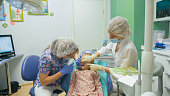 Child with a mother at a dentist's reception. The girl lies in the chair, behind her mother. The doctor works with an assistant. Procedure for drilling a tooth. Setting a temporary seal.