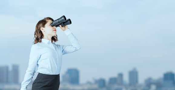 Young businesswoman using binoculars in front of the city.