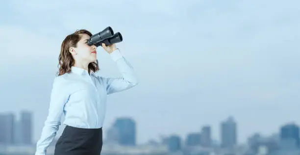 Photo of Young businesswoman using binoculars in front of the city.