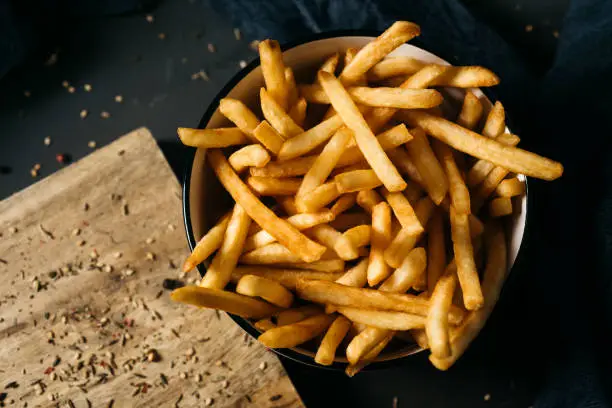 high angle shot of some appetizing french fries served in a white ceramic bowl, placed on a dark gray rustic wooden table