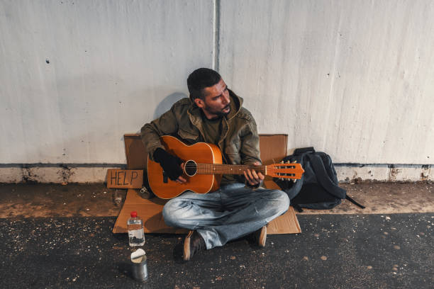 Photo of young beggar or Homeless dirty man singing and playing guitar in city with donate bowl, paper cardboard with help text to ask for donation. Poverty in town. Unemployed person, Street Performer with alcohol problem holding acoustic guitar in subway.