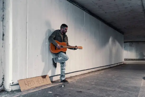 Photo of young beggar or Homeless dirty man standing, singing and playing guitar on stair of modern city with donate bowl, paper cardboard with need help text to ask for donation. Poverty in town. Unemployed person, Street Performer with alcohol problem holding acoustic guitar in subway.