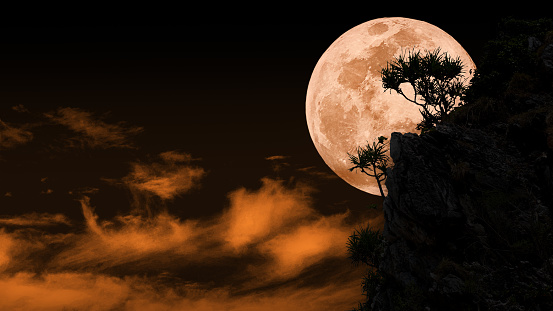 Silhouette of full blue blood moon behind rocky cliff with background of orange sky