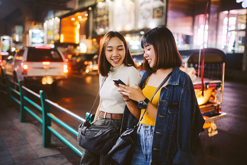 Vintage toned image of two young, Asian women, walking together, chatting, near Siam station on Bangkok Sukhumvit line. They’re wearing casual street style clothing, enjoying an afternoon together, texting on their cellphones.