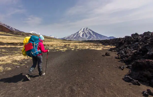 man in red jacket with backpack going on black volcanic slag with view on ridge of snowy volcanoes of Kamchatka with blue clouded sky