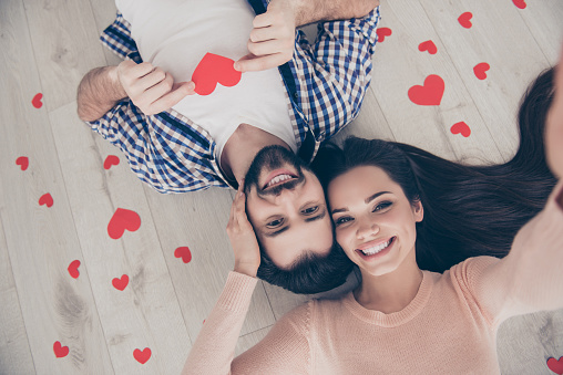 Self portrait of sweet cute couple lying head to head on the floor with small red hearts around shooting selfie on front camera, having fun leisure at home indoor