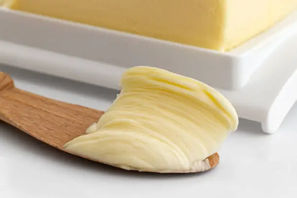 Detail of butter spread on wooden knife next to a plastic butter dish on white background.