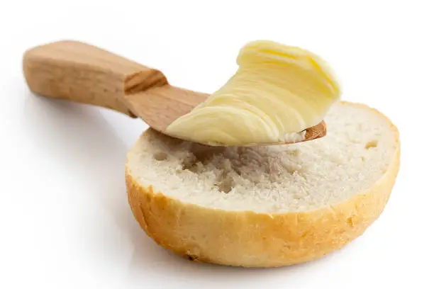 Butter on wooden knife resting on half of crusty bread roll isolated on white.