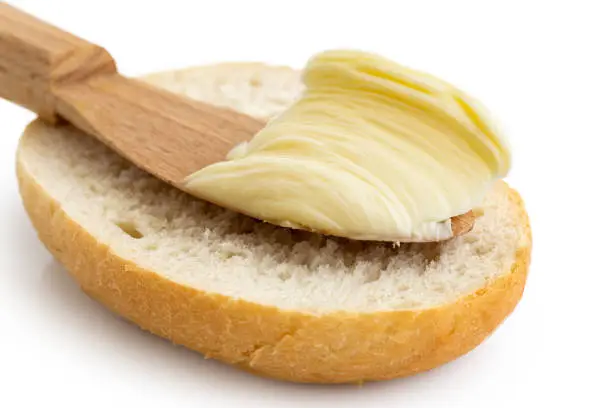 Detail of butter on wooden knife resting on half of crusty bread roll on white background.