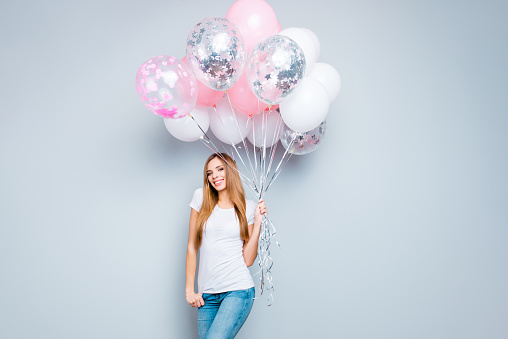 Portrait of trendy nice girl with long hair in casual outfit having pink and white air balloons in hand looking at camera isolated on grey background