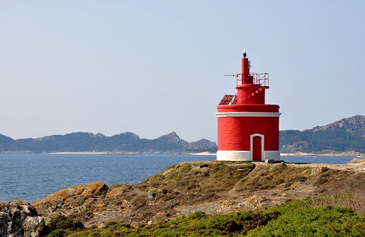 Red and white lighthouse in coastline with Cies Islands in the background in Vigo Bay, Spain