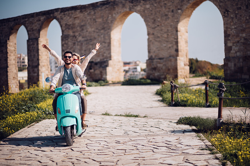 Young tourists couple on summer holidays in Europe riding retro scooter with arms outstretched