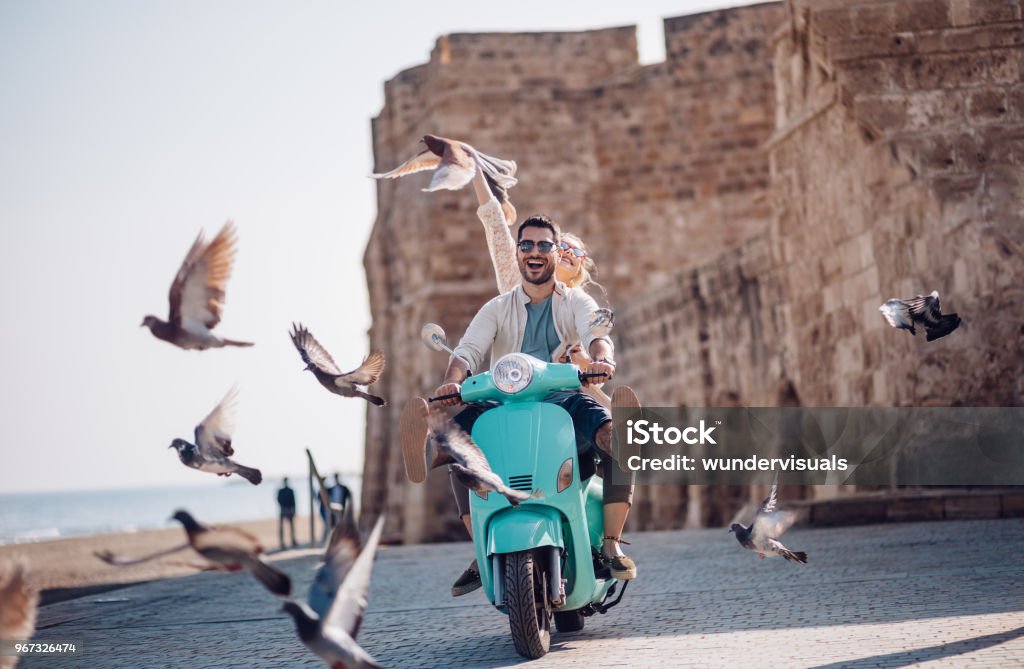 Young couple having fun riding scooter in old European town Tourist friends on summer holidays in Europe having fun riding retro scooter by the sea Travel Stock Photo