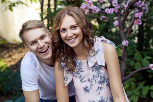 Cheerful young couple spends quality time together in a park on a sunny day. The woman playfully embraces the man from behind while he holds a vibrant bouquet of fresh flowers. Both are dressed in stylish autumn clothes, capturing a joyful and romantic moment in an outdoor urban setting.