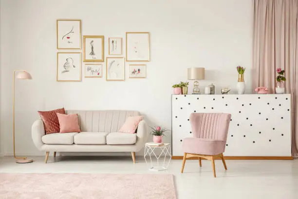Pink chair next to table and sofa in living room interior with posters and gold lamp