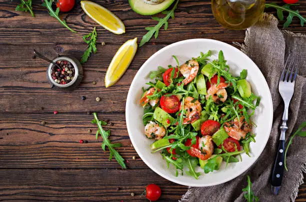Photo of Fresh salad bowl with shrimp, tomato, avocado and arugula on wooden background close up. Healthy food. Clean eating. Top view. Flat lay.