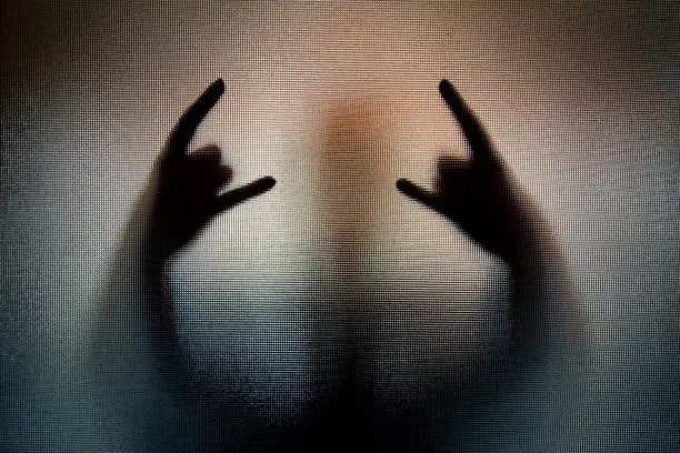 Color image depicting the shadow silhouette of a person performing the heavy metal sign of the horns hand gesture, behind frosted glass. The person's body is distorted and spooky, giving an atmospheric, mood to the image. Lots of room for copy space.