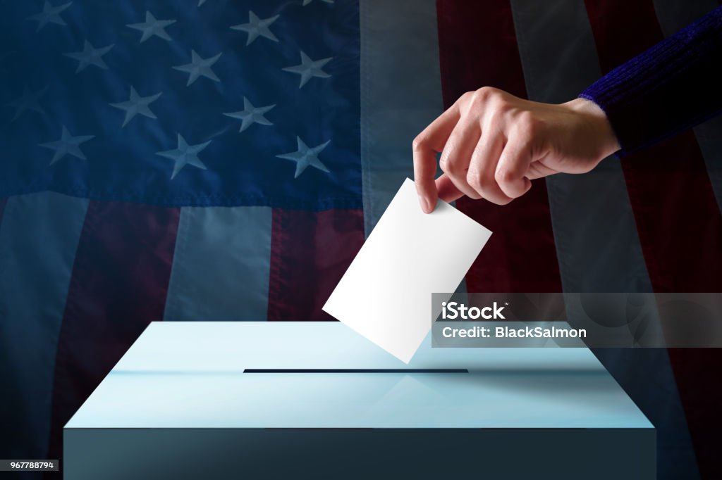 Election in America Concept. Hand Dropping a Ballot Card into the Vote Box, Flag of United States as background Voting Stock Photo