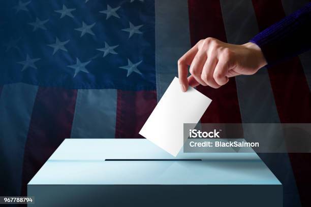 Election In America Concept Hand Dropping A Ballot Card Into The Vote Box Flag Of United States As Background Stock Photo - Download Image Now
