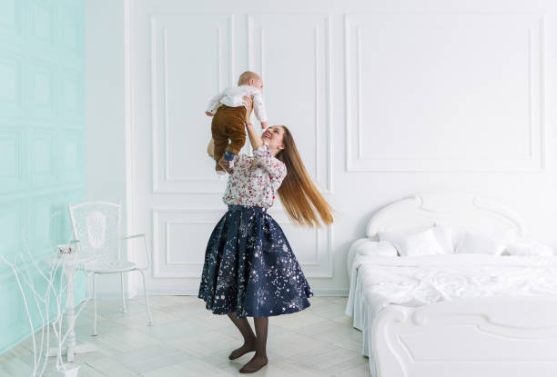 Young attractive mother plays with her baby at home Young attractive mother plays with her baby at home, wearing dress, standing and holding her baby daughter high, playful interaction and communication baby boy dancing with his mommy stock pictures, royalty-free photos & images