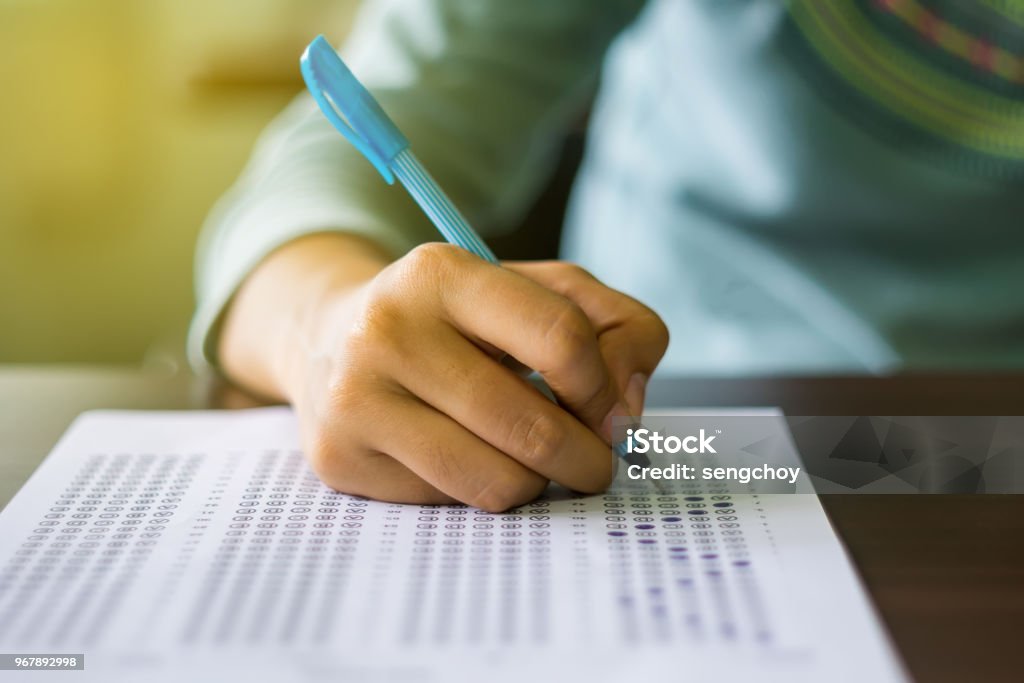 Close up of high school or university student holding a pen writing on answer sheet paper in examination room. College students answering multiple choice questions test in testing room in university. Close up of high school or university student holding a pen writing on answer sheet paper in the examination room. College students answering multiple choice questions test in the testing room in university. Educational Exam Stock Photo