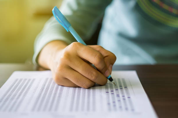 Close up of high school or university student holding a pen writing on answer sheet paper in examination room. College students answering multiple choice questions test in testing room in university. stock photo
