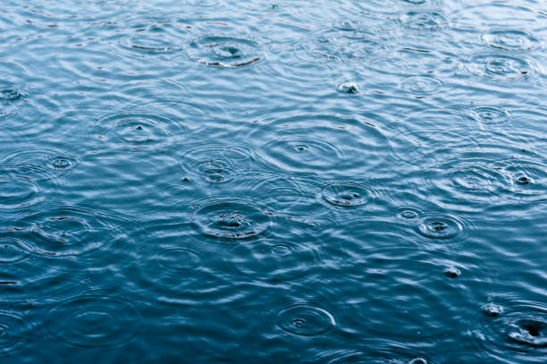 la lluvia cae sobre la superficie del agua en un charco con graduado sombra de sombra negra y el reflejo del cielo azul - puddle condensation water drop fotografías e imágenes de stock