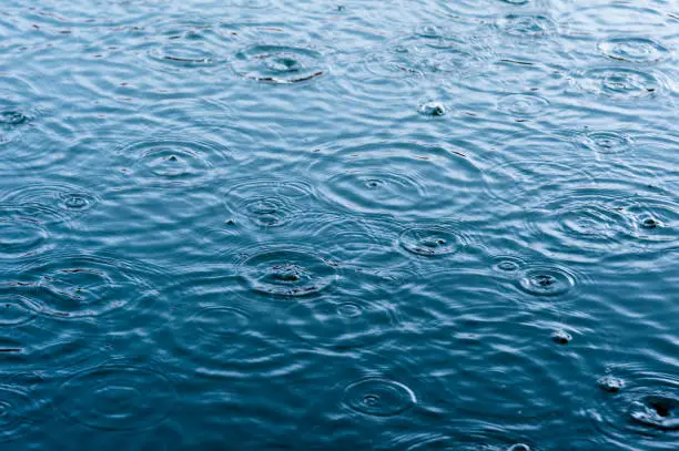 Photo of rain drops on the surface of water in a puddle with graduated shade of black shadow and reflection of blue sky