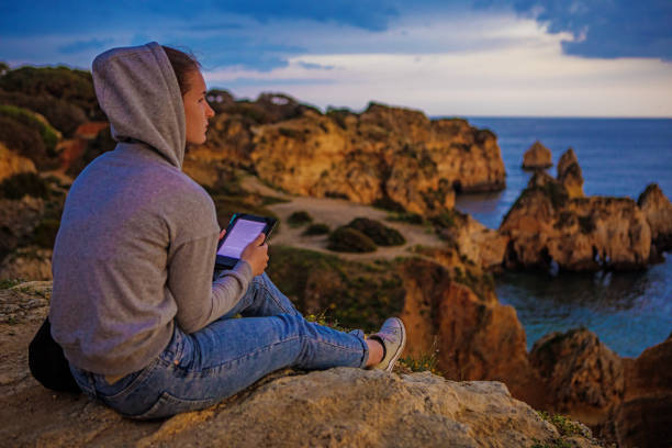 Young brunette female reading her e-book on a cliff in Alvor Portugal in late summer sun