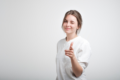 Positive cheerful young caucasian woman wearing white casual T-shirt blinking her eyes and smiling pointing at camera with index fingers. Facial emotion. Succees in life or studies
