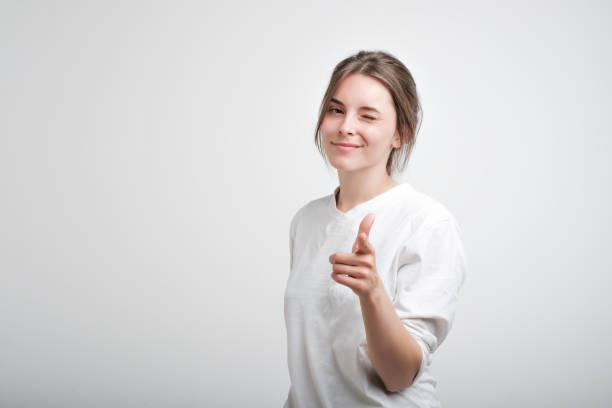 Positive cheerful young caucasian woman wearing white casual T-shirt blinking her eyes and smiling pointing at camera stock photo