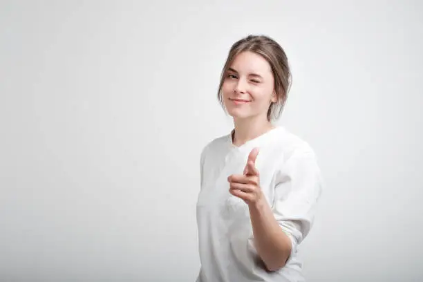 Photo of Positive cheerful young caucasian woman wearing white casual T-shirt blinking her eyes and smiling pointing at camera