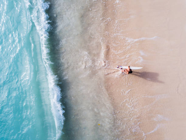 Surfer woman walking out of the ocean holding a surfboard (from above) stock photo