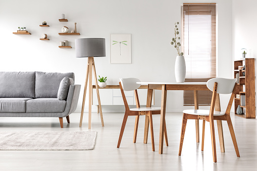 Wooden chairs at table in bright open space interior with lamp next to grey couch. Real photo