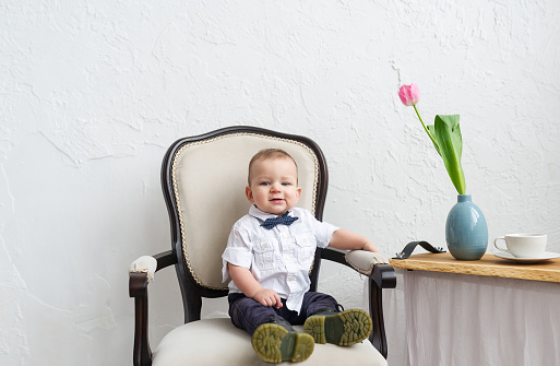 Fashionable baby boy sitting in the armchair at white room. Horizontal portrait