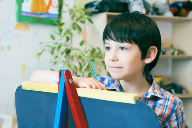istock Child standingnext to the easel. Kid boy learn paint by brush in class school. Kindergarten interior on background. Boy is getting ready to become an artist 968261094