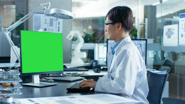 istock Asian Scientist Sitting at His Desk Works on a Personal Computer with Mock-up Green Screen. In the Background Computer Science Research Laboratory with Robotic Arm Model. 968289620