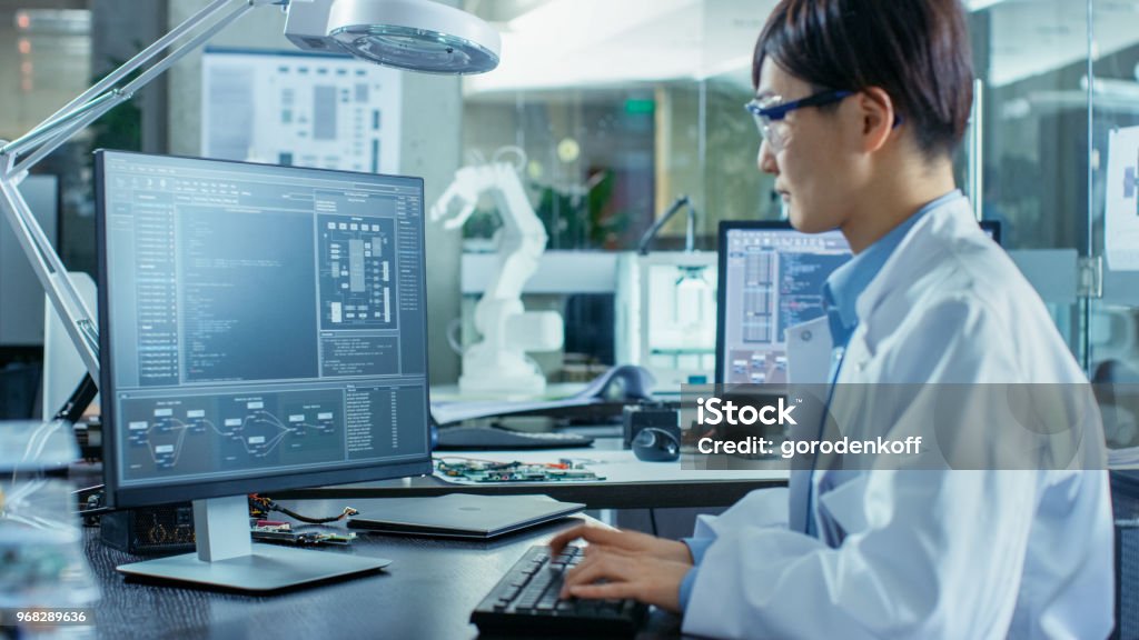 Asian Scientist Sitting at His Desk Doing Sophisticated Coding and Programming on His Desktop Computer. In the Background Computer Science Research Laboratory with Robotic Arm Model. Research Stock Photo