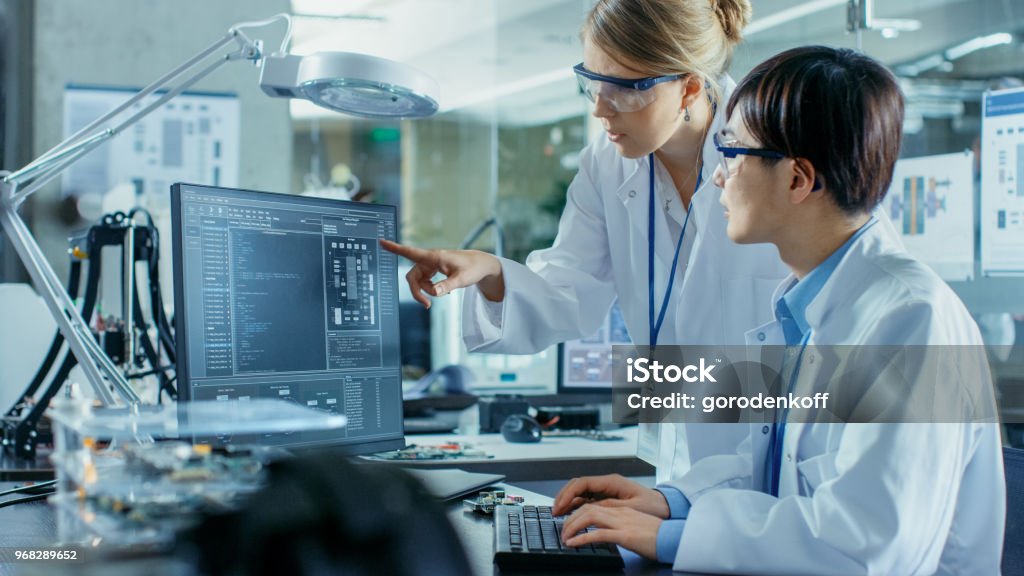 Asian Scientist Sitting at His Desk Consults Senior Engineer about Sophisticated Coding and Programming. In the Background Computer Science Research Laboratory with Robotic Arm Model. Laboratory Stock Photo