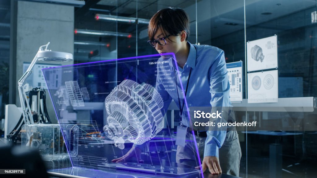 Asian Neural Network Engineer Uses Modern Computer With Transparent Holographic Display. Monitor Shows Visualization of a Mechanical Part. Shot in Modern Glass and Concrete Office. Engineer Stock Photo