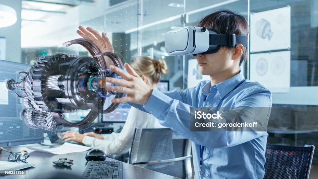 Computer Science Engineer wearing Virtual Reality Headset Works with 3D Model Hologram Visualization, Makes Gestures. In the Background Engineering Bureau with Busy Coworkers. Virtual Reality Stock Photo