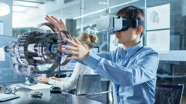 Photo of Computer Science Engineer wearing Virtual Reality Headset Works with 3D Model Hologram Visualization, Makes Gestures. In the Background Engineering Bureau with Busy Coworkers.