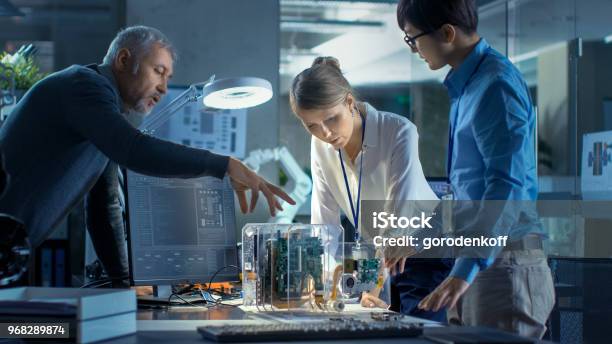 Team Of Computer Engineers Lean On The Desk And Choose Printed Circuit Boards To Work With Computer Shows Programming In Progress In The Background Technologically Advanced Scientific Research Center Stock Photo - Download Image Now