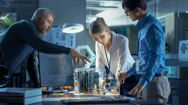 equipe de engenheiros de computador inclinar-se sobre a mesa e escolher placas de circuito impresso para trabalho com computador mostra a programação em andamento. no fundo tecnologicamente avançado centro de pesquisa científica. - leaning - fotografias e filmes do acervo