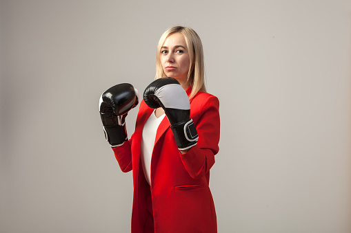 Young beautiful white blond girl in a bright red strict suit with a jacket and white blouse standing in a pose with boxing gloves on a white isolated background