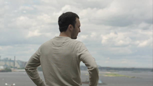 Young man is looking into the distance to the river on a cloudy day. Professional shot on BMCC RAW with high dynamic range. You can use it e.g in your commercial video,music video.