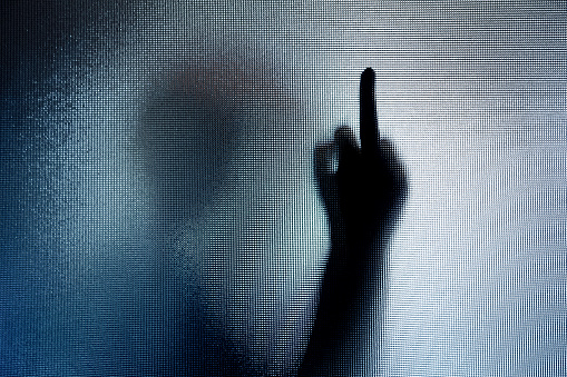 Color image depicting the silhouette of a person performing the rude swearing hand gesture of holding up the middle finger behind frosted glass. The person's body is distorted and spooky, giving an atmospheric, mood to the image. Lots of room for copy space.