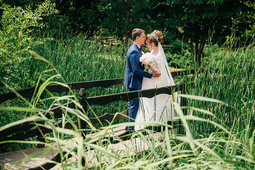 A man in a blue suit and a girl in a wedding dress and a bouquet of flowers stand on a bridge and gently embrace. They want to get married today, and are very happy about it