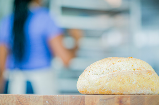 A loaf of bread is sitting on a table inside a bakery. The baker is preparing ingredients in the background.