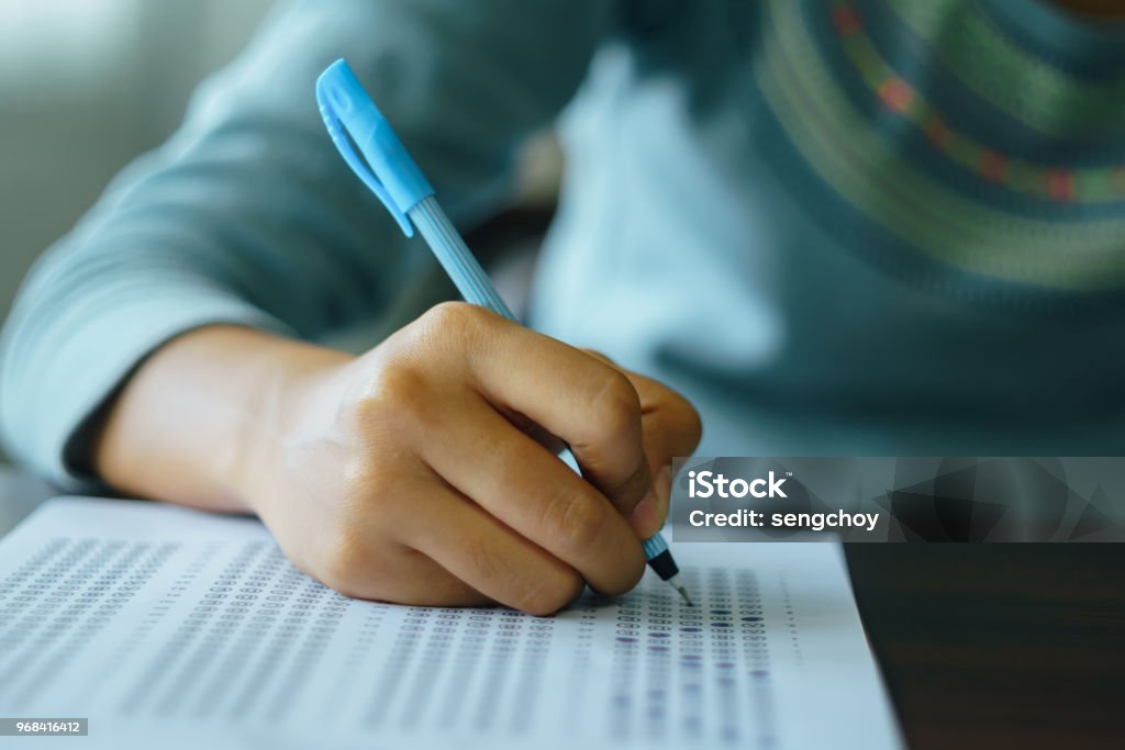 College student write on exam paper Close up of high school or university student holding a pen writing on answer sheet paper in the examination room. College students answering multiple choice questions test in the testing room in university. Educational Exam Stock Photo